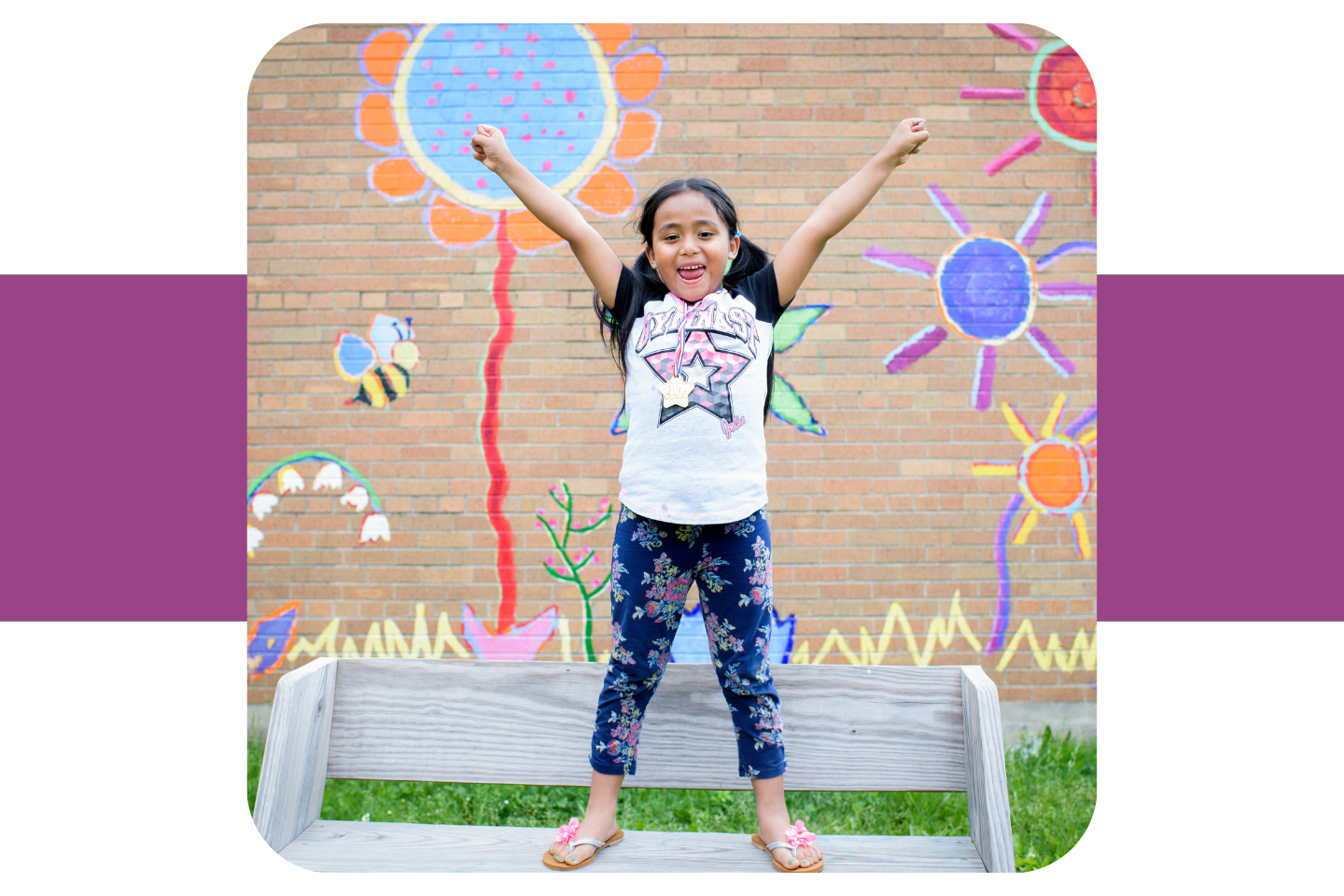 A small girls stands smiling in on a bench with her arms in the air.